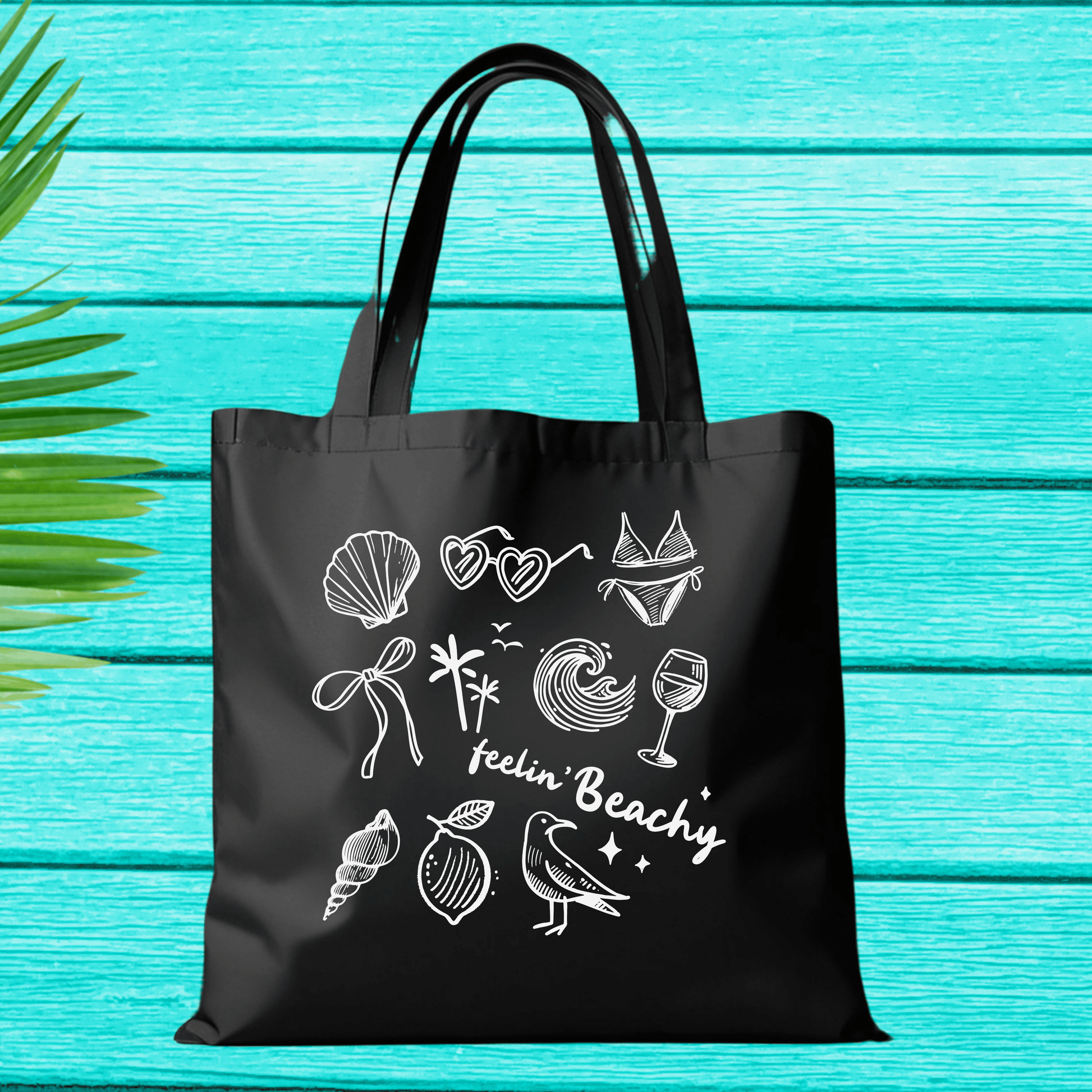 Black Canvas Tote Bag with "Feelin' Beachy" Design featuring seashells, palm tree, waves, and bird on large reusable bag.
