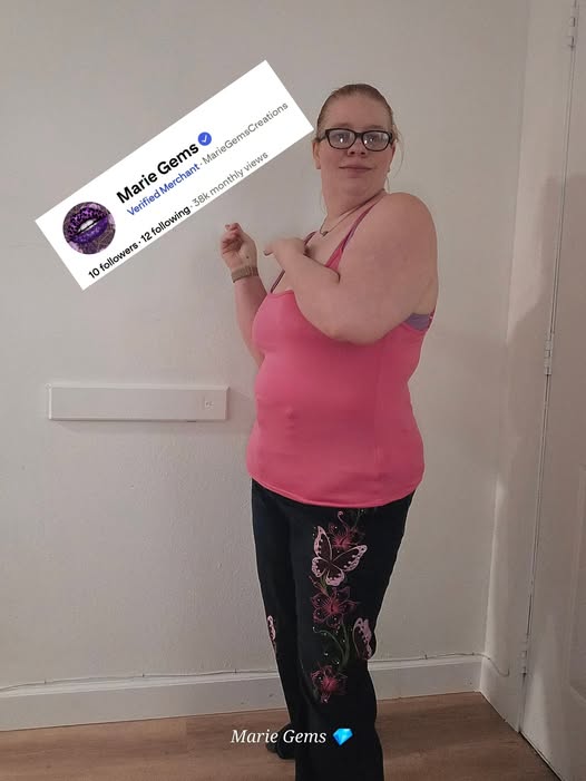 Marie wearing a pink top and black pants with butterfly designs, standing indoors. pointing to her Verified Merchant Pinterest Profile !