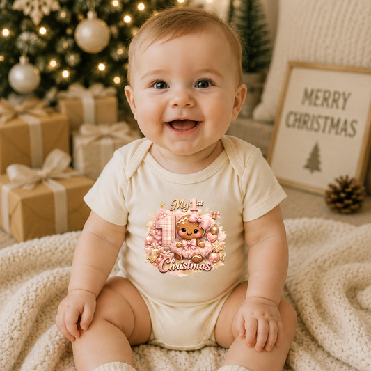 Smiling baby girl wearing a Baby Girl First Christmas bodysuit with a pink gingerbread Christmas design while sitting in front of a decorated Christmas tree.