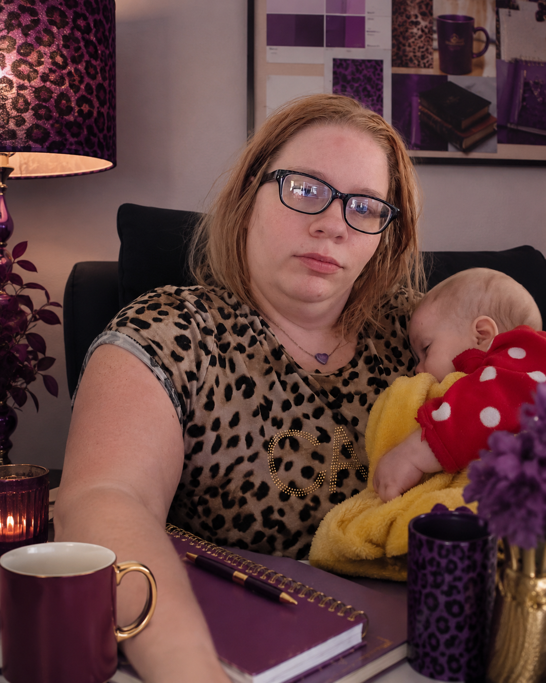 Realistic mom entrepreneur working from home while holding a sleeping baby in a cozy purple and cheetah print inspired workspace.