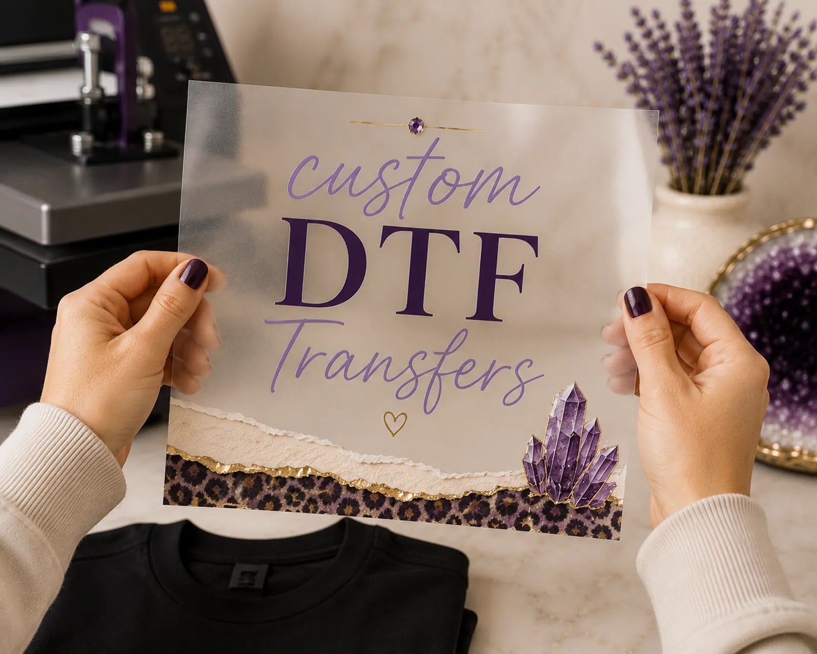 Hands holding a custom DTF transfer sheet with 'custom DTF Transfers' script and serif text over a heat press and folded black shirt, styled with amethyst crystals - Marie Gems