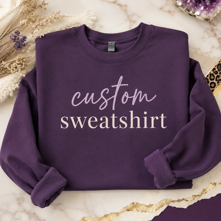 Folded purple custom sweatshirt with 'custom sweatshirt' script and serif text design, styled on marble background with dried flowers and amethyst crystal - Marie Gems