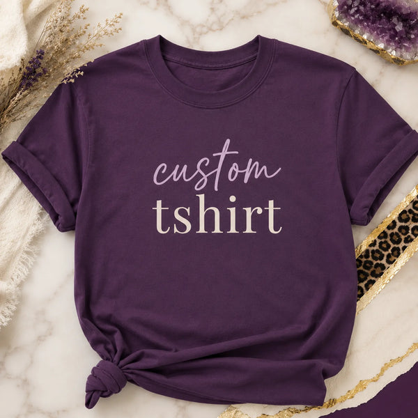 Folded purple custom t-shirt with 'custom tshirt' script and serif text design, styled on marble background with dried flowers and amethyst crystal - Marie Gems