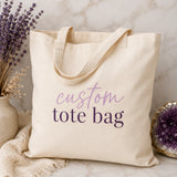 Natural canvas custom tote bag with 'custom tote bag' script and serif text design, styled with dried lavender and amethyst crystal - Marie Gems