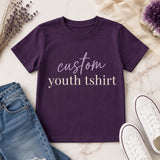 Folded purple custom youth t-shirt with 'custom youth tshirt' script and serif text design, styled on marble background with dried flowers and amethyst crystal - Marie Gems
