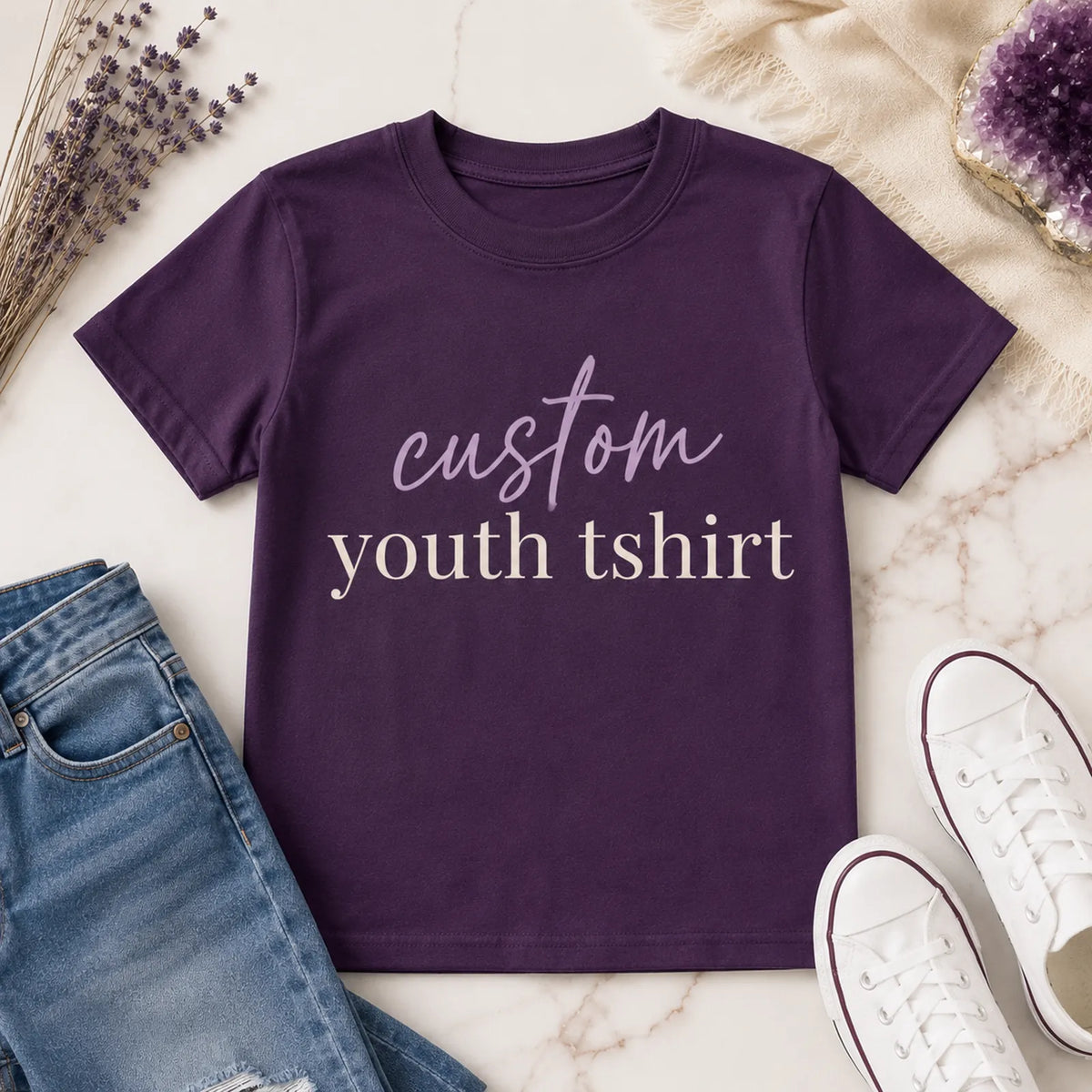 Folded purple custom youth t-shirt with 'custom youth tshirt' script and serif text design, styled on marble background with dried flowers and amethyst crystal - Marie Gems
