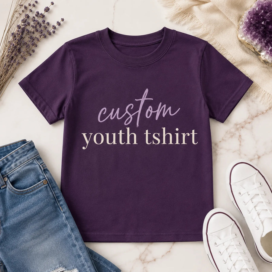 Folded purple custom youth t-shirt with 'custom youth tshirt' script and serif text design, styled on marble background with dried flowers and amethyst crystal - Marie Gems