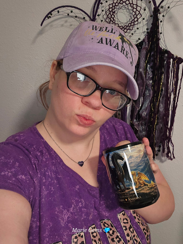 Marie of Marie Gems wearing a purple western graphic tee and baseball cap well fuck aware from fourth wing while holding a custom coffee cup of tarin and andana from fourth wing book in a cozy lifestyle branding photo