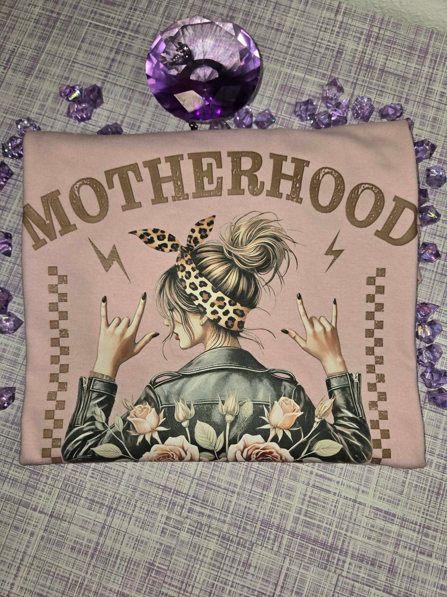 Front design of a light pink motherhood graphic tee featuring floral roses, leopard print accents, and a rocker-inspired mom design displayed in a styled flat lay product photo. 💜