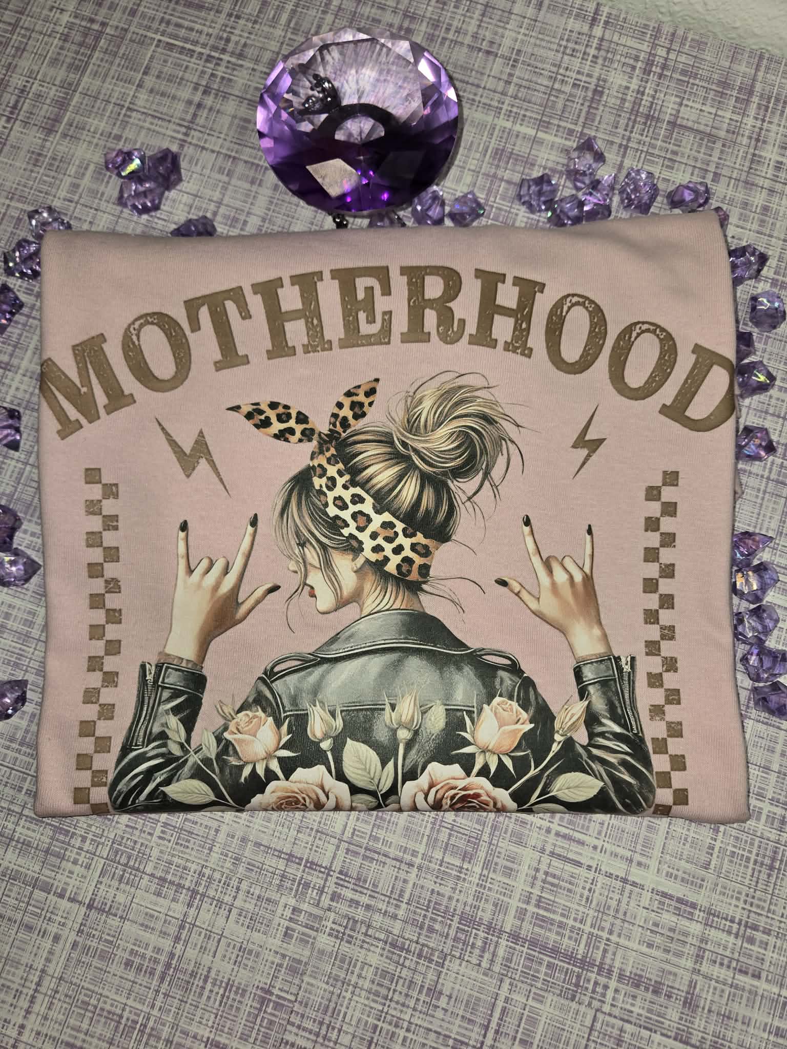 Front design of a light pink motherhood graphic tee featuring floral roses, leopard print accents, and a rocker-inspired mom design displayed in a styled flat lay product photo. 💜