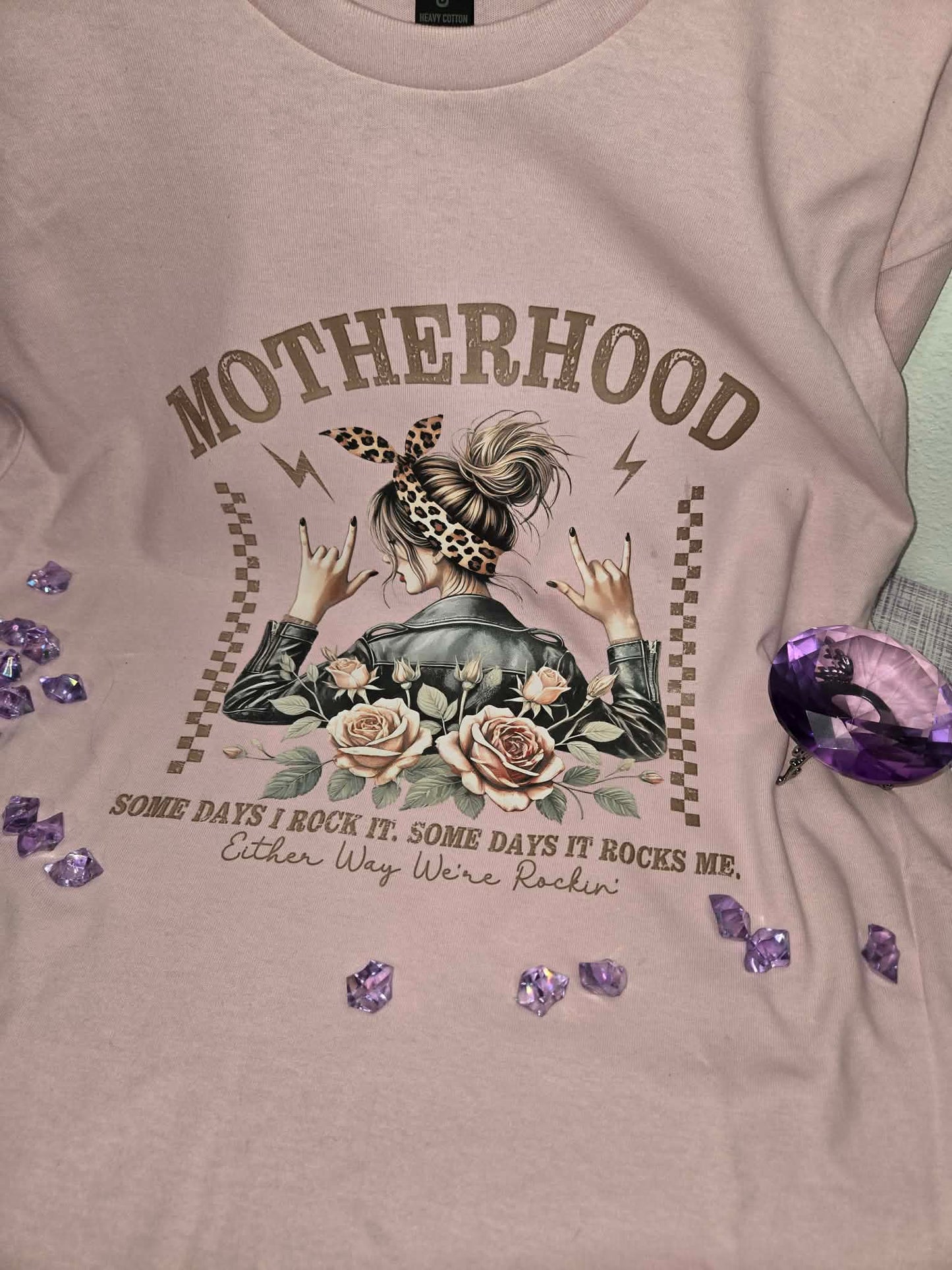 Light pink motherhood graphic tee featuring floral roses, leopard print accents, and the quote Some Days I Rock It Some Days It Rocks Me in a styled flat lay product photo.
