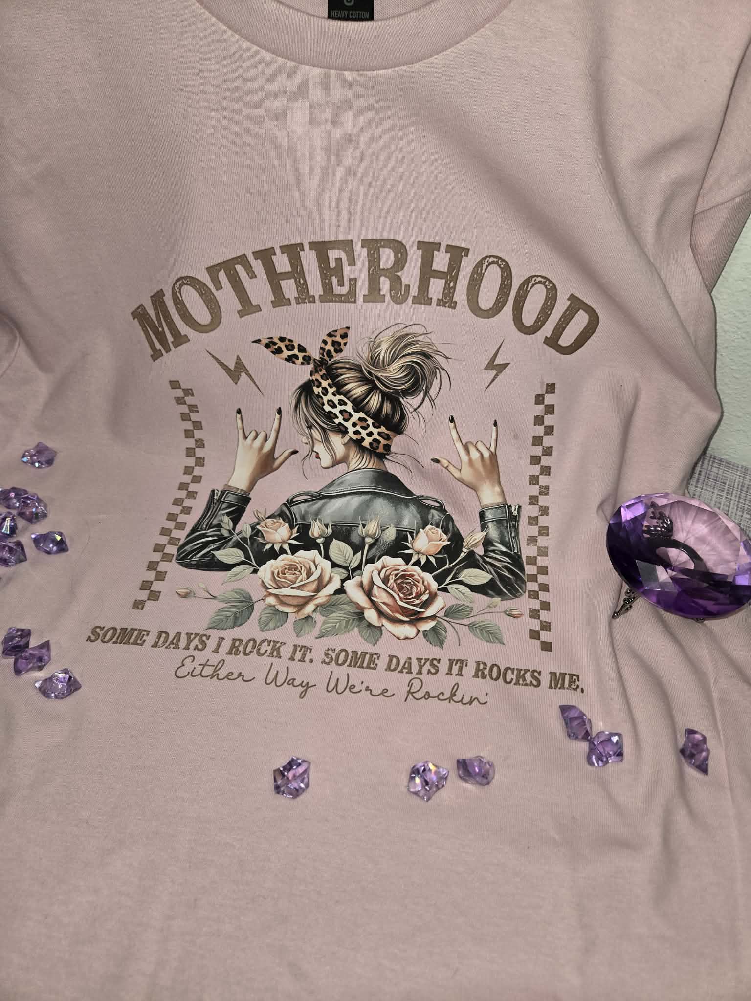 Light pink motherhood graphic tee featuring floral roses, leopard print accents, and the quote Some Days I Rock It Some Days It Rocks Me in a styled flat lay product photo.