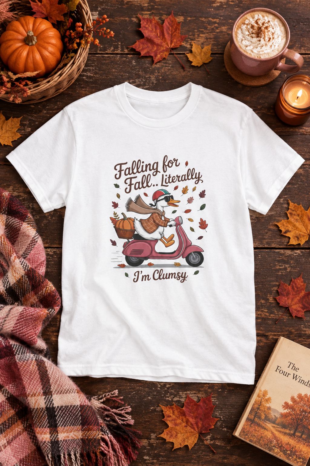 Falling for Fall Literally I'm Clumsy t-shirt in white with goose on a scooter graphic, styled flat lay with a pumpkin basket, plaid scarf, autumn leaves and a latte