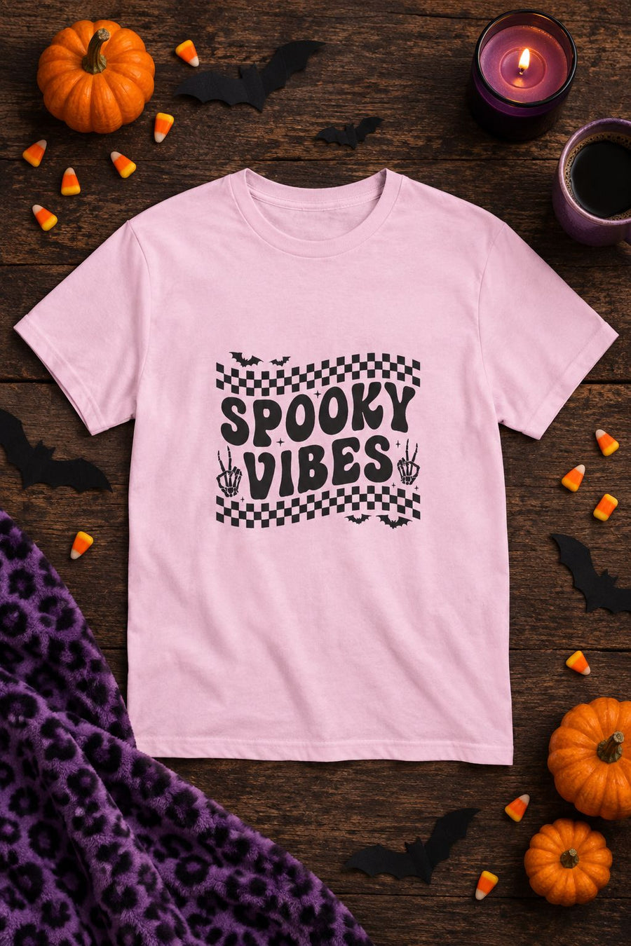 Spooky Vibes Retro Checkerboard t-shirt in light pink with checkerboard Spooky Vibes graphic, styled flat lay with pumpkins, candy corn, bats and a purple candle