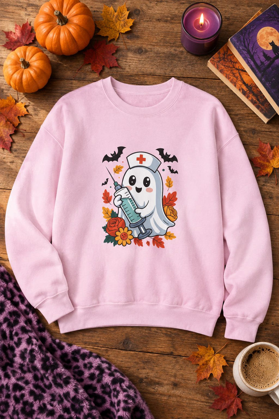 Nurse Ghost Halloween crewneck sweatshirt in light pink with cute nurse ghost graphic, styled flat lay with pumpkins, a purple candle, books and a stethoscope