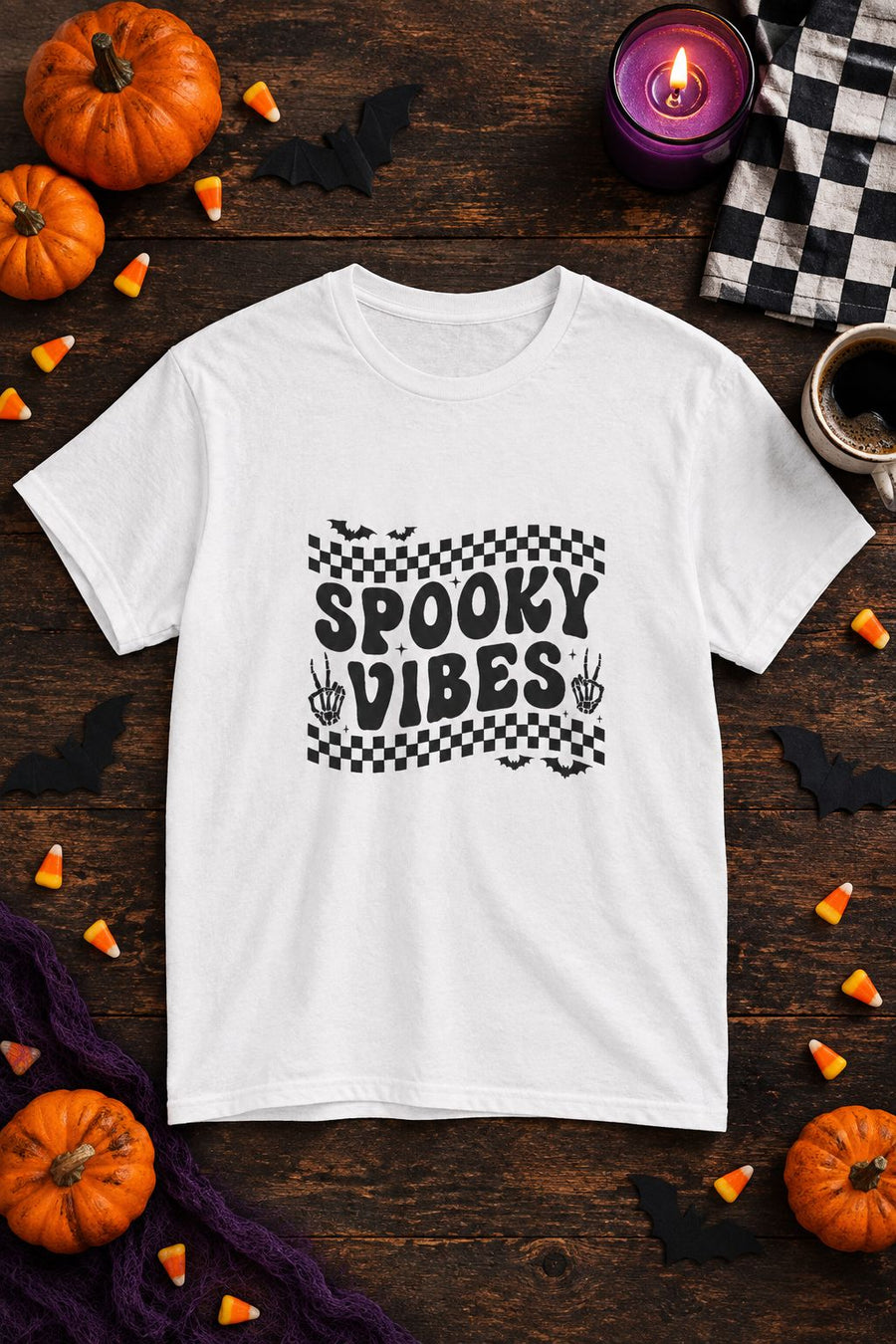 Spooky Vibes Retro Checkerboard t-shirt in white with checkerboard Spooky Vibes graphic, styled flat lay with pumpkins, candy corn, bats and a purple candle