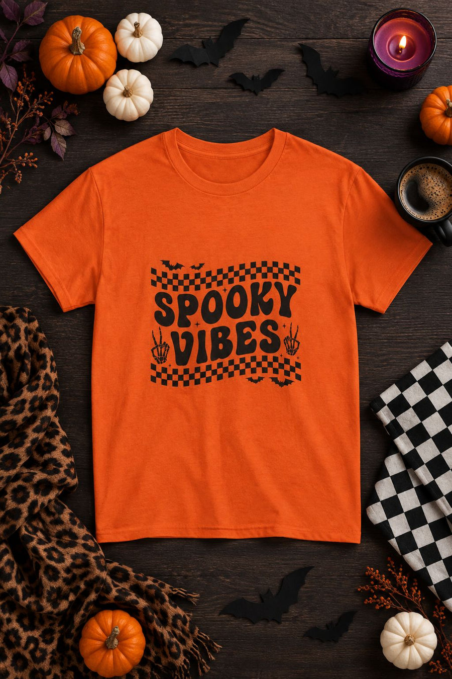Spooky Vibes Retro Checkerboard t-shirt in orange with checkerboard Spooky Vibes graphic, styled flat lay with pumpkins, candy corn, bats and a purple candle