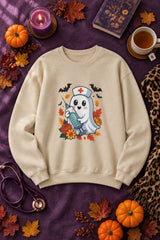 Nurse Ghost Halloween crewneck sweatshirt in sand with cute nurse ghost graphic, styled flat lay with pumpkins, a purple candle, books and a stethoscope