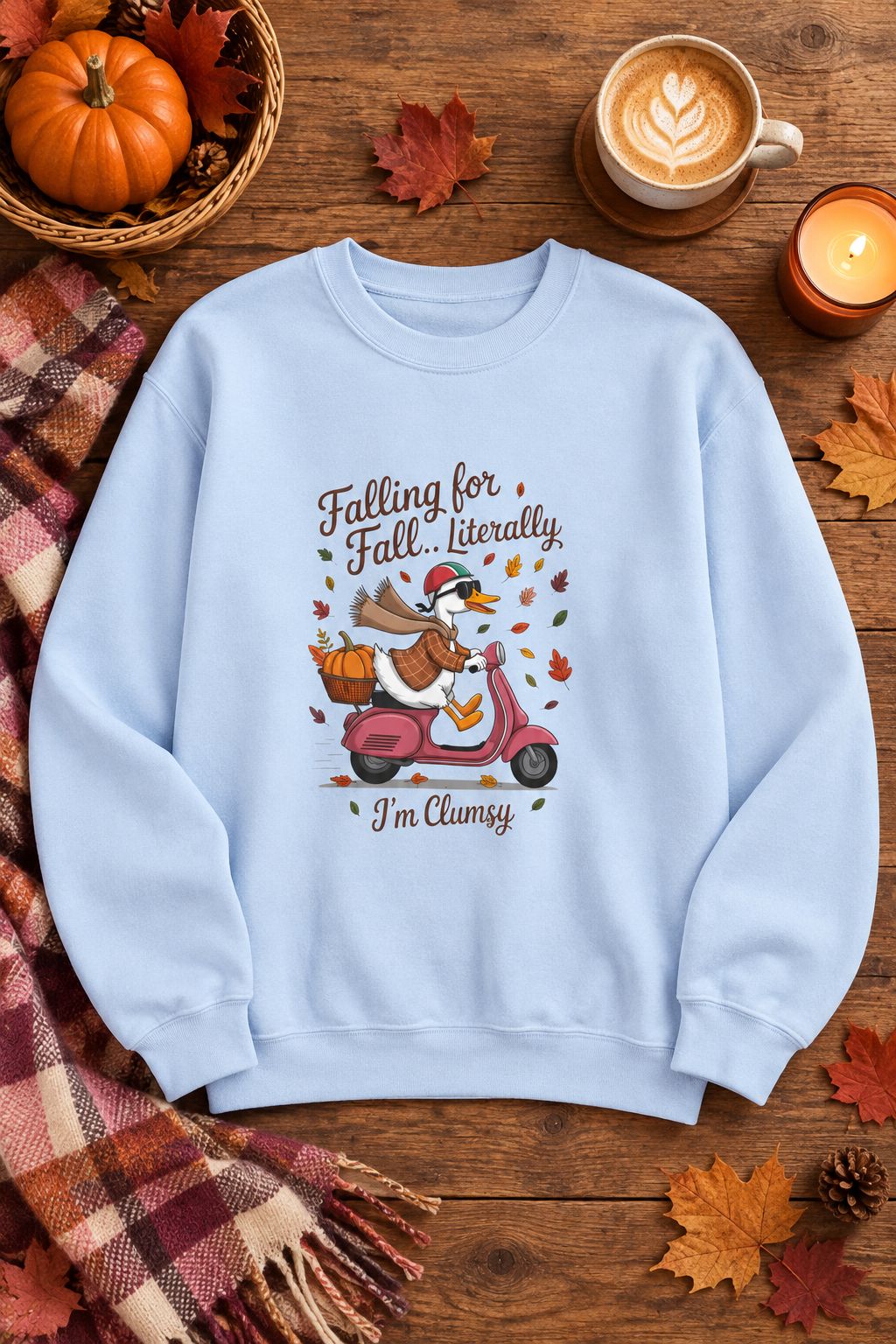 Falling for Fall Literally I'm Clumsy crewneck sweatshirt in light blue with goose on a scooter graphic, styled flat lay with a pumpkin basket, plaid scarf, autumn leaves and a latte