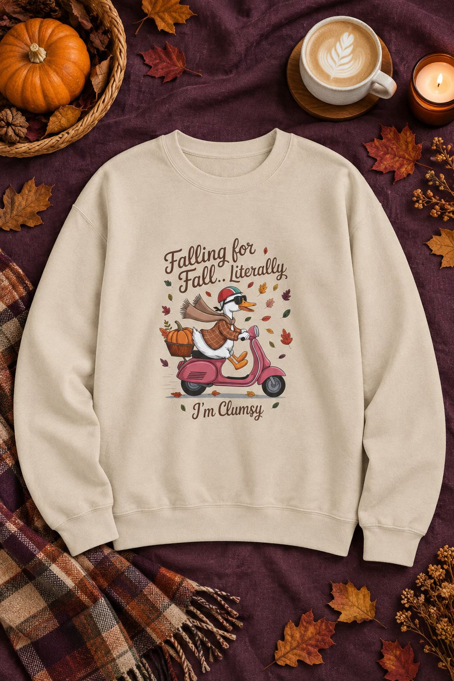 Falling for Fall Literally I'm Clumsy crewneck sweatshirt in sand with goose on a scooter graphic, styled flat lay with a pumpkin basket, plaid scarf, autumn leaves and a latte