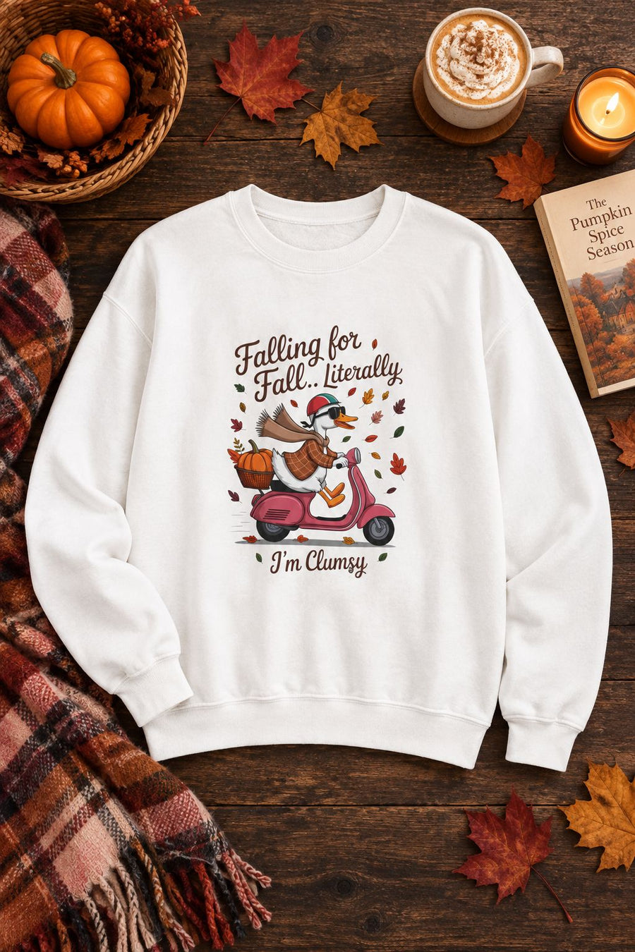 Falling for Fall Literally I'm Clumsy crewneck sweatshirt in white with goose on a scooter graphic, styled flat lay with a pumpkin basket, plaid scarf, autumn leaves and a latte