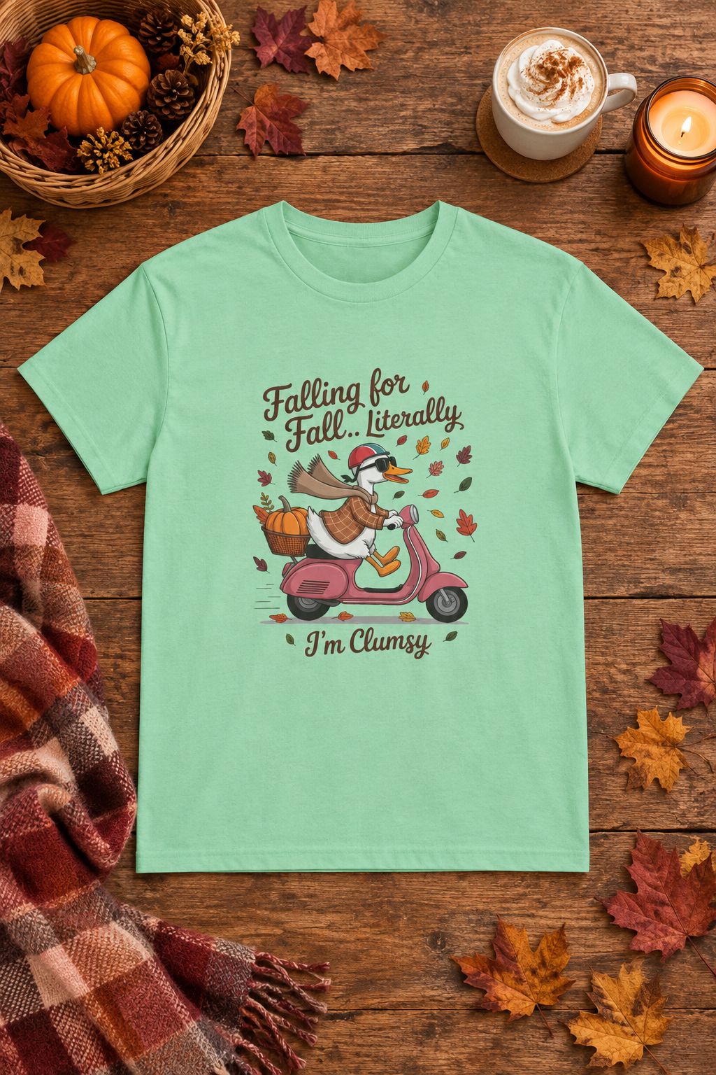 Falling for Fall Literally I'm Clumsy t-shirt in mint green with goose on a scooter graphic, styled flat lay with a pumpkin basket, plaid scarf, autumn leaves and a latte