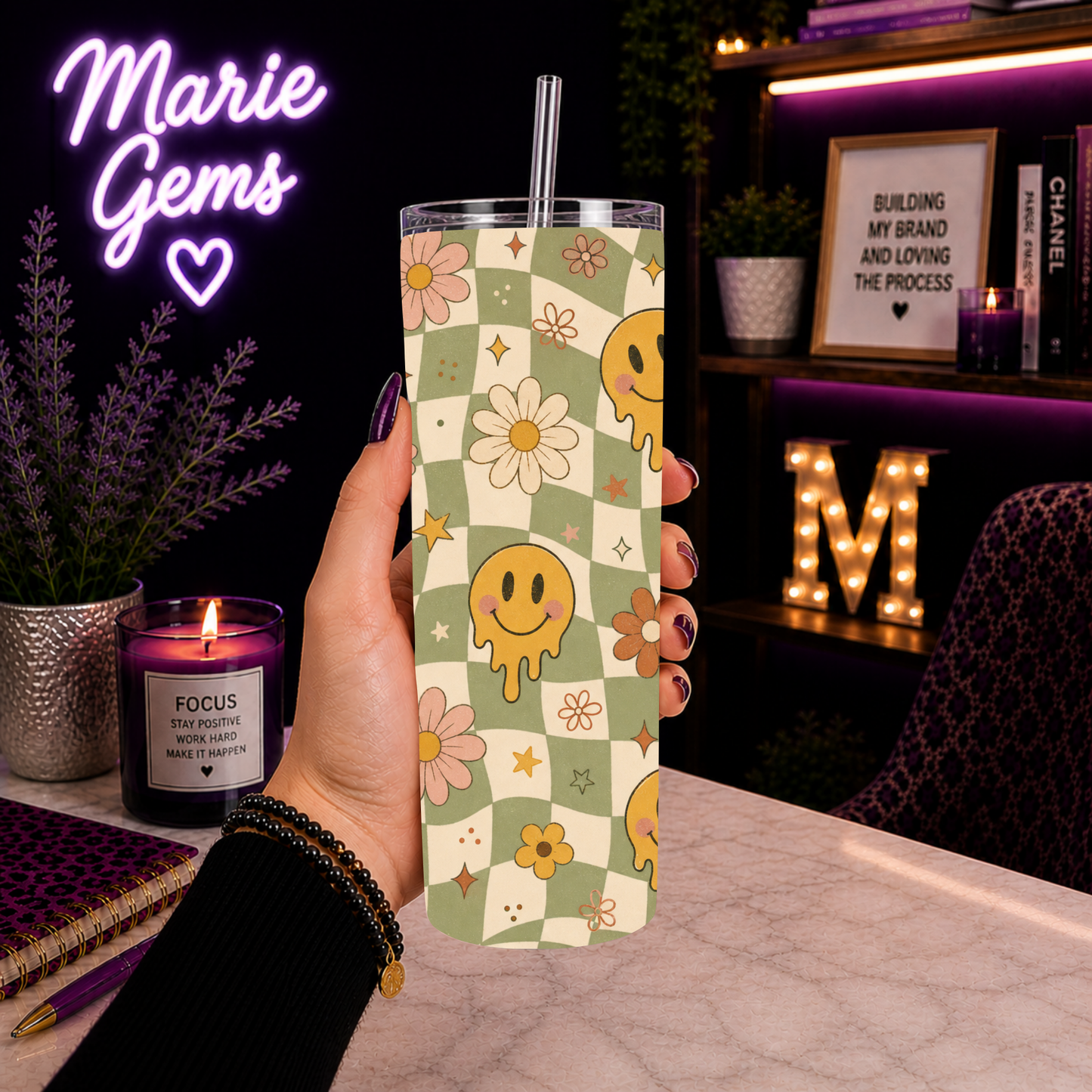 A hand holds a clear tumbler with a metal straw, featuring a pastel green and cream checkered pattern decorated with yellow smiley faces, pink and yellow flowers, and small stars. The background is a cozy, dimly lit room with a purple neon 'Marie Gems' sign, a lit candle, and shelves with books and decor.